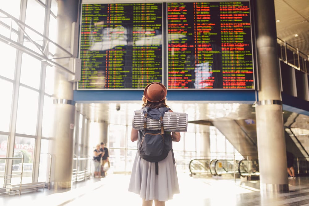 Theme travel public transport. young woman standing with back in dress and hat behind backpack and camping equipment for sleeping, insulating mat looks schedule on scoreboard airport station young woman standing in an airport