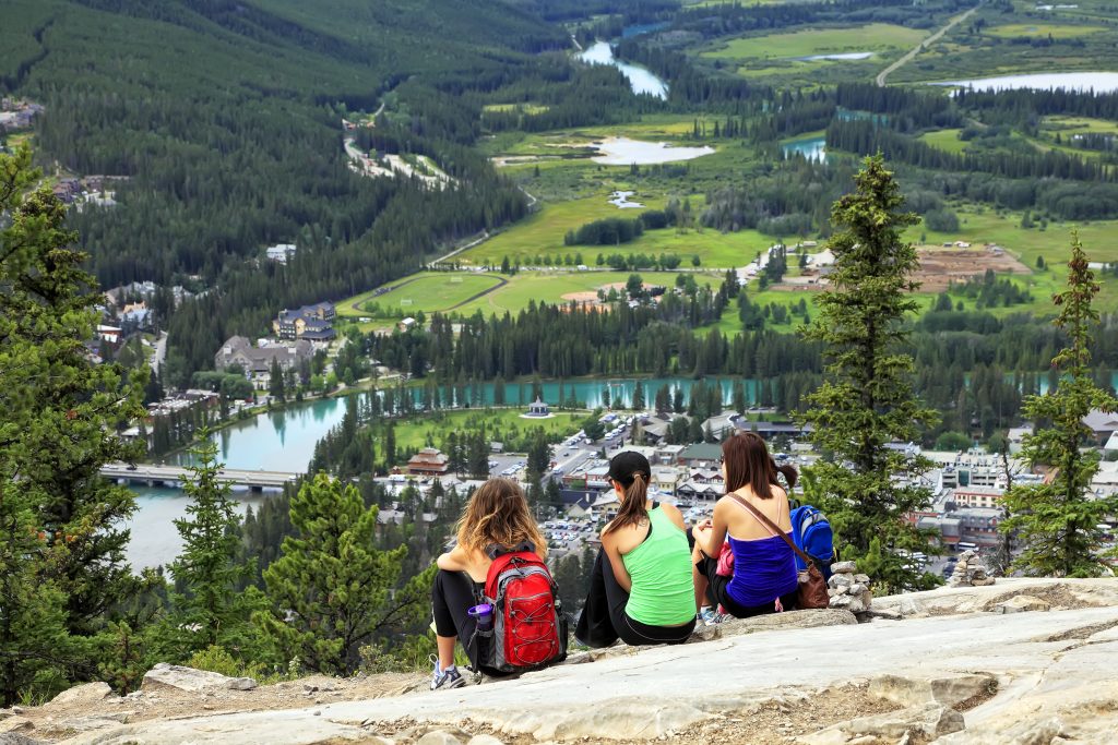 Three girls-tourists sitting on a grief and looking at a panorama of a small town Banff in a Bow river valley